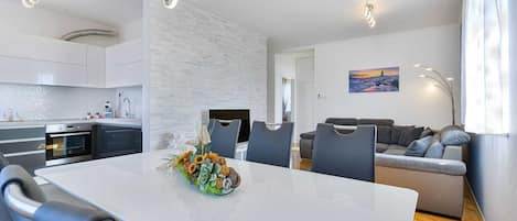 A modern kitchen with built-in appliances, a dining area with a white table and chairs, and a living room with a sofa and a painting on the wall.