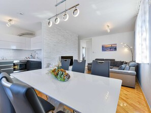 Une cuisine moderne dotée d’appareils électroménagers intégrés, un coin repas avec une table et des chaises blanches, et un salon avec un canapé et un tableau au mur.