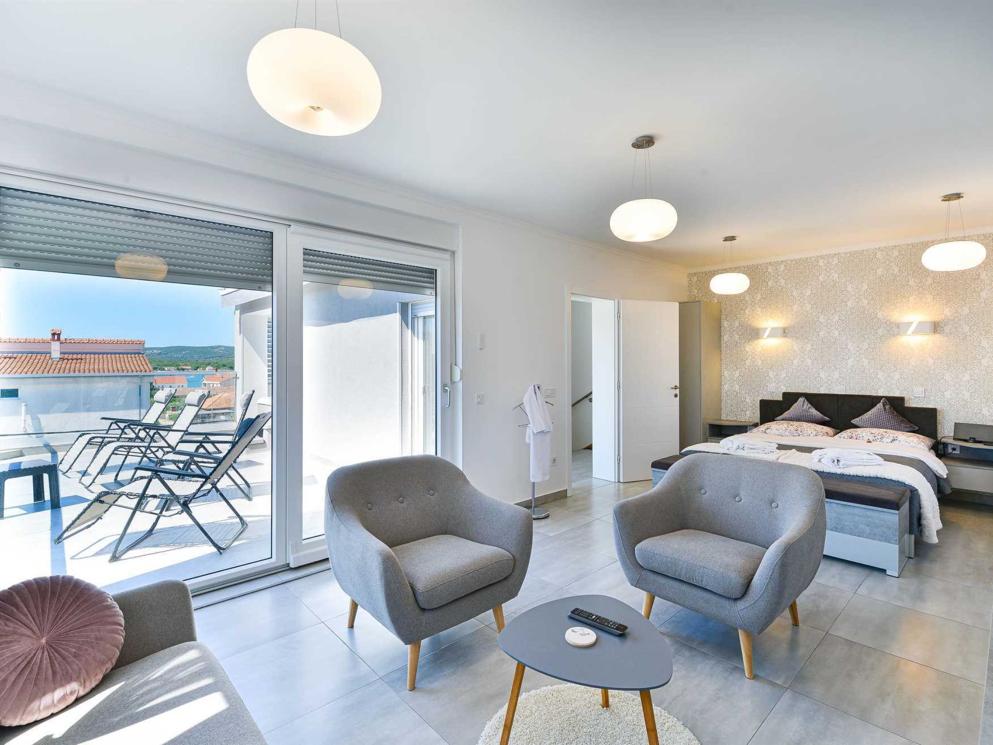 Ein modernes Hotelzimmer mit Balkon, zwei Sesseln, einem runden Couchtisch, einem Bett mit Kopfteil und einem großen Fenster.