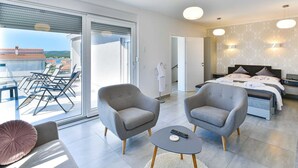 Een moderne hotelkamer met balkon, twee fauteuils, een ronde salontafel, een bed met hoofdeinde en een groot raam.