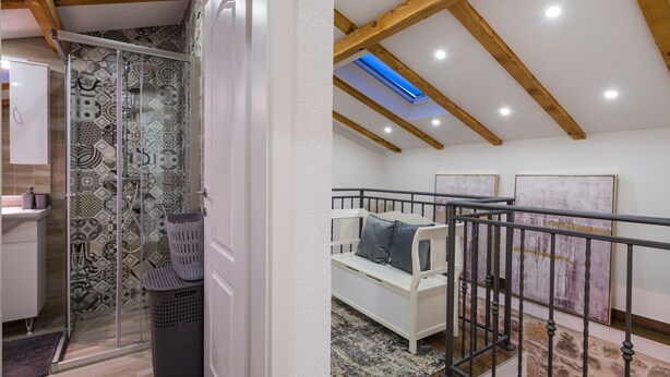 Ein Zimmer mit hohen Decken, Holzbalken, einer Glasdusche, gemusterten Fliesen, einer weißen Bank mit Kissen und einer Steinwand.