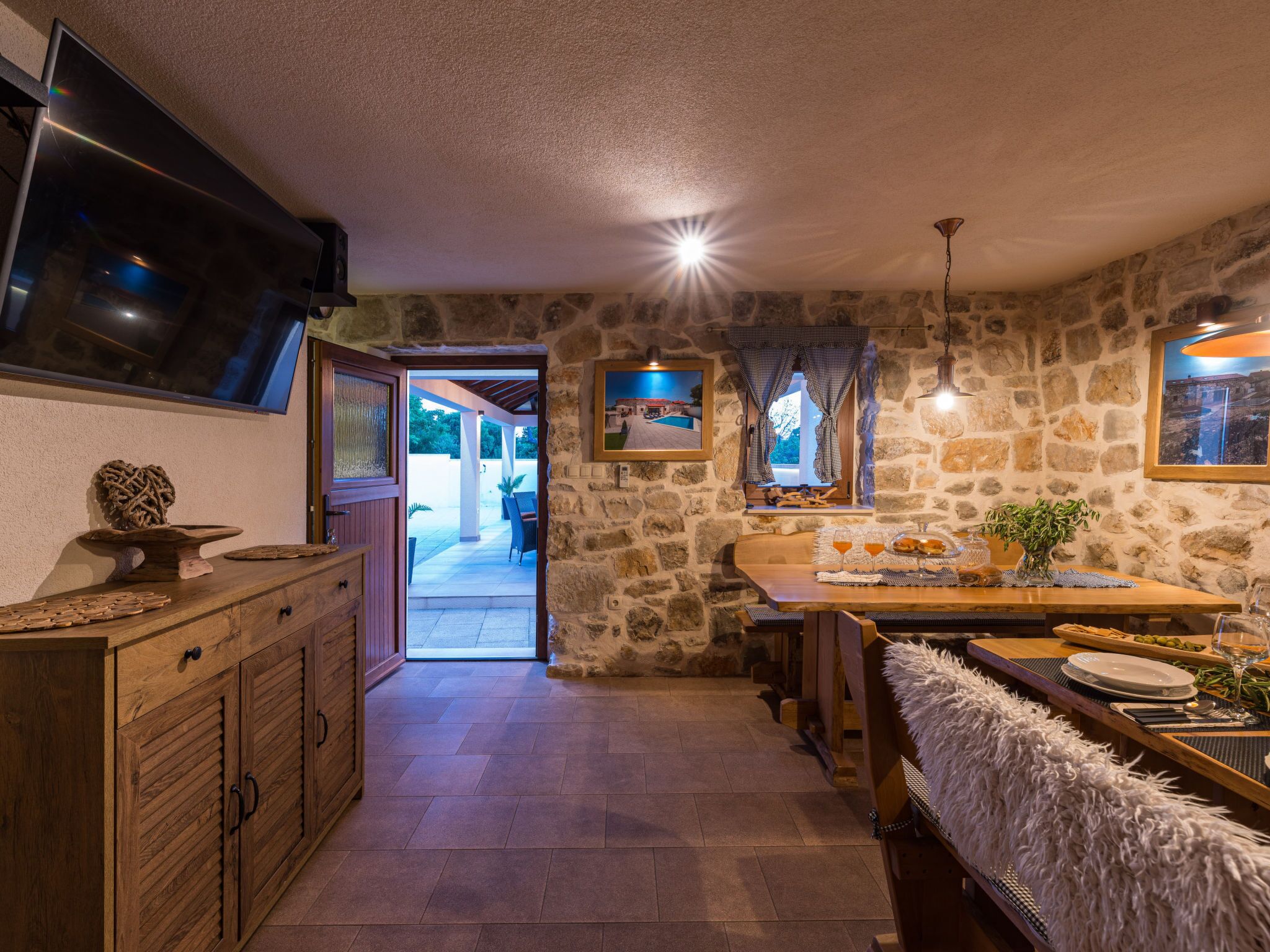 Un intérieur rustique avec des murs en pierre, une table à manger en bois dressée pour deux personnes et un téléviseur fixé au mur.