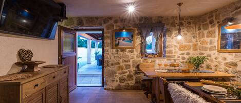 Un intérieur rustique avec des murs en pierre, une table à manger en bois dressée pour deux personnes et un téléviseur fixé au mur.