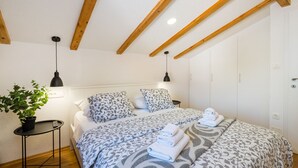 A bedroom with a sloped ceiling, wooden beams, a bed with patterned bedding, a bedside table with a plant, and a hanging lamp.