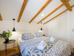 A bedroom with a sloped ceiling, wooden beams, a bed with patterned bedding, a bedside table with a plant, and a hanging lamp.