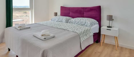 A neatly made bed with a white and purple headboard, a bedside table with a lamp, and a view of the outdoors through a window.
