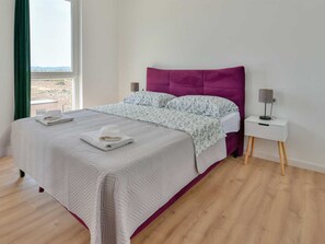 A neatly made bed with a white and purple headboard, a bedside table with a lamp, and a view of the outdoors through a window. - Villa Silente with pool and sauna by Interhome (Debeljak)