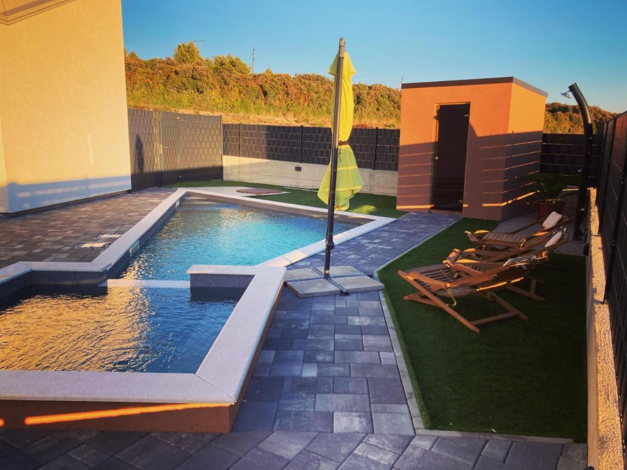 A swimming pool with a sunken area, surrounded by a patio with lounge chairs and a small building.