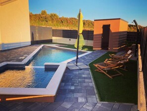 Ein Schwimmbecken mit einer Vertiefung, umgeben von einer Terrasse mit Liegestühlen und einem kleinen Gebäude.