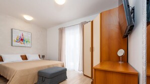 Una camera d'albergo con un letto, una scrivania, una sedia, un poggiapiedi, una TV e una finestra con tende.