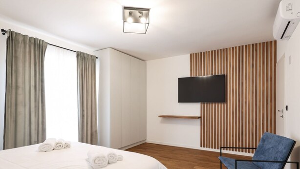 Ein Hotelzimmer mit einem Bett, einem Sessel, einem an einer Holzvertäfelung befestigten Fernseher und einer Deckenleuchte.