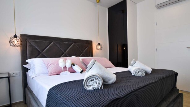 Ein ordentlich bezogenes Bett mit Kissen, einem zusammengerollten Handtuch und einer Decke. Es befinden sich zwei Wandleuchten an der Wand.