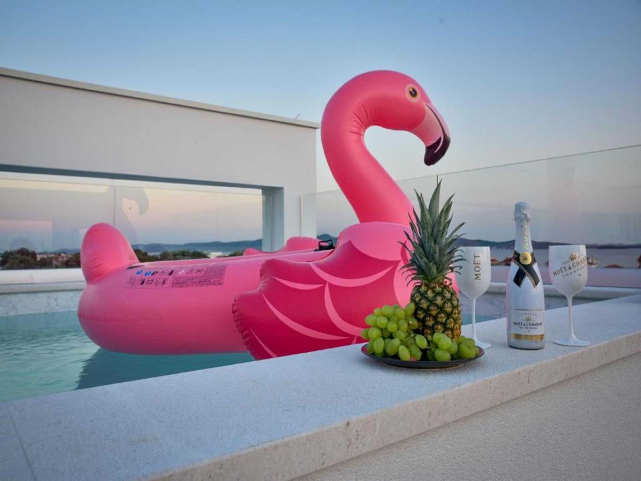 A poolside setting with a pink flamingo float, a pineapple, a bottle of champagne, and two glasses.