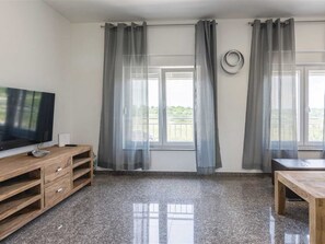 Ein Zimmer mit Fernseher auf einem Holzboden, einem kleinen Tisch und zwei Fenstern mit grauen Vorhängen.