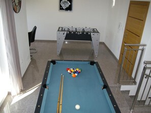 Ein Billardtisch mit Kugeln und Spielstangen, ein Kicker, ein Stuhl und eine Tür.