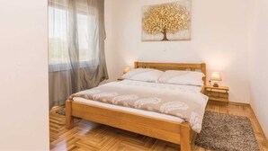 A bedroom with a wooden bed, a nightstand with a lamp, a window with curtains, and a wall-mounted tree artwork. - Vacation home  by Interhome (Crno)