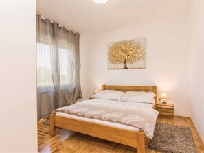 Ein Schlafzimmer mit einem hölzernen Bett, einem Nachttisch mit Lampe, einem Fenster mit Vorhängen und einem an der Wand hängenden Baum-Kunstwerk.