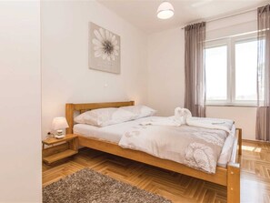Ein Einzelbett mit Holzrahmen, weiße Bettwäsche, eine Nachttischlampe, ein kleiner Teppich, ein Fenster mit Vorhängen und ein Blumenbild an der Wand.