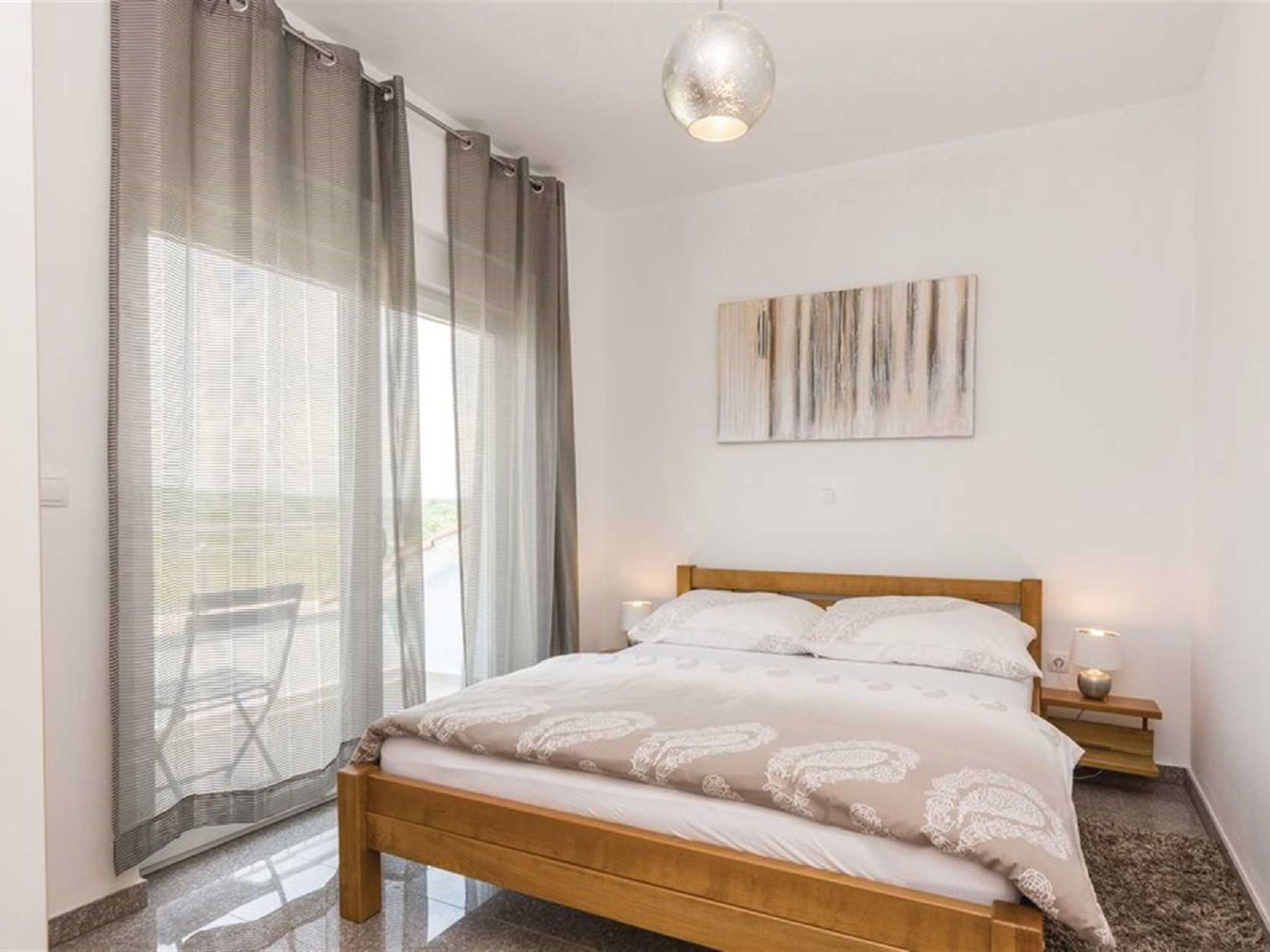 A bedroom with a bed, a nightstand, a lamp, a window with curtains, and a painting on the wall.