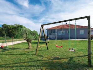 Ein Spielplatz mit Schaukeln und einem umzäunten Bereich, ein Baum und ein Haus mit rotem Dach im Hintergrund.