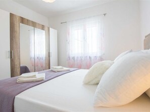 Ein Schlafzimmer mit Bett, Kissen, einem Stapel Handtücher, einem Kleiderschrank und einem Fenster mit durchsichtigen Vorhängen.