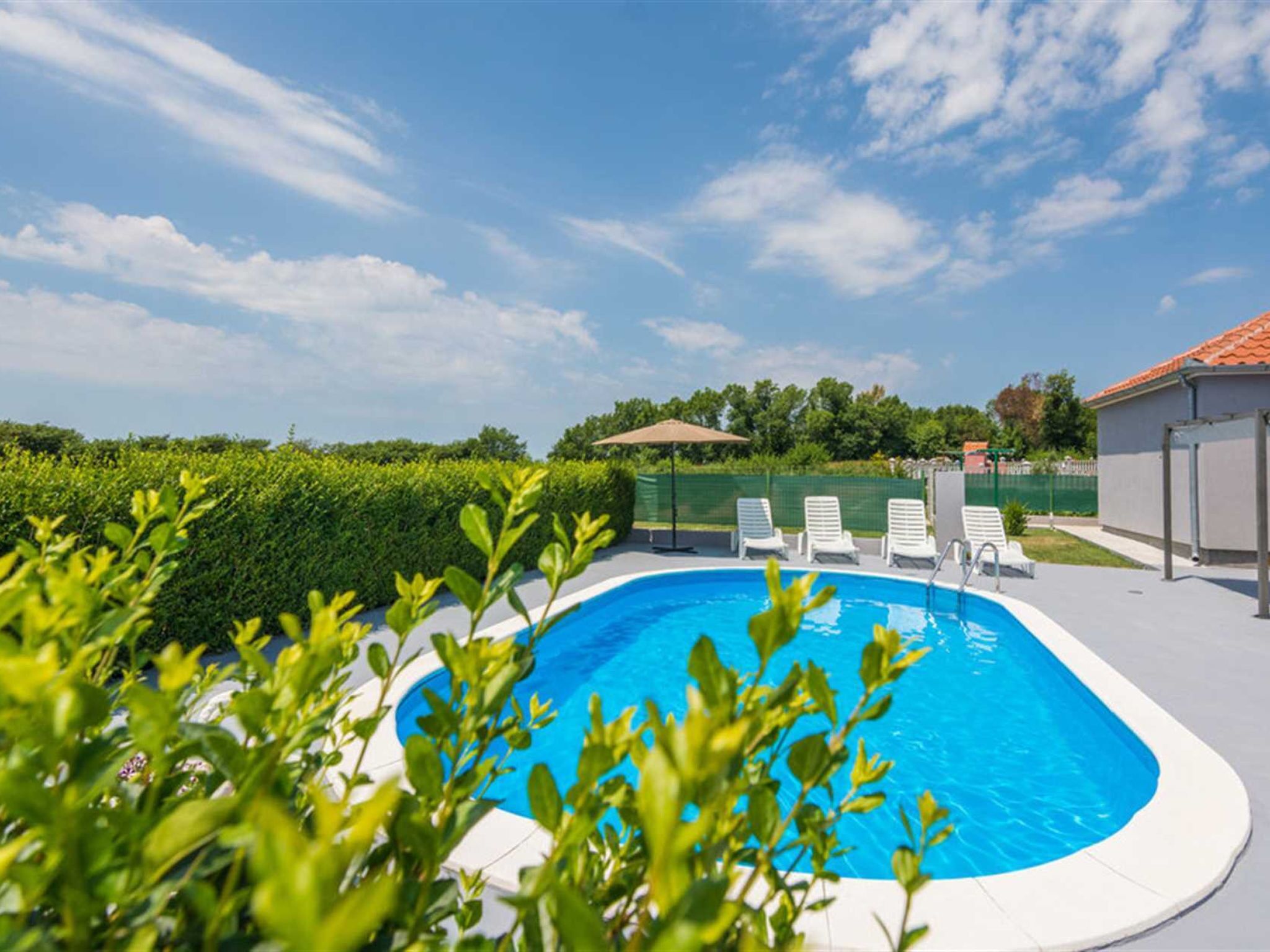 A swimming pool with a clear blue water, surrounded by greenery and a fenced area with lounge chairs.