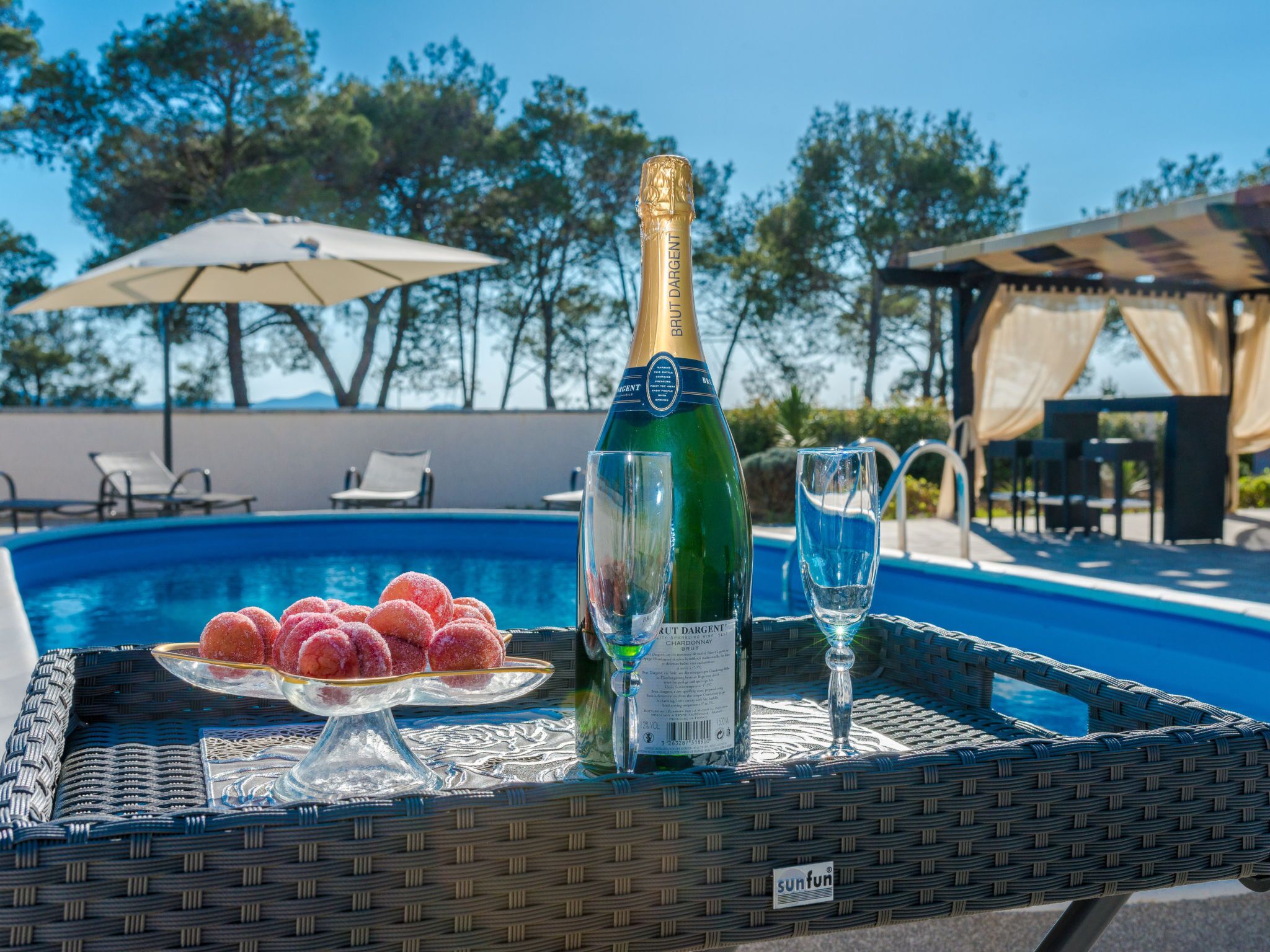 A bottle of Brut Dargent champagne, two glasses, and a tray with sugared doughnuts by a poolside.