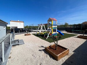 Ein Spielplatz mit Rutsche und Schaukeln, umgeben von Kiesflächen und einem eingezäunten Grundstück.