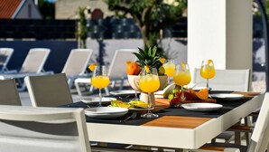 Un espace de restauration en plein air avec une table dressée pour deux personnes, agrémentée d’un panier de fruits, de verres de jus d’orange et d’une variété de fruits.