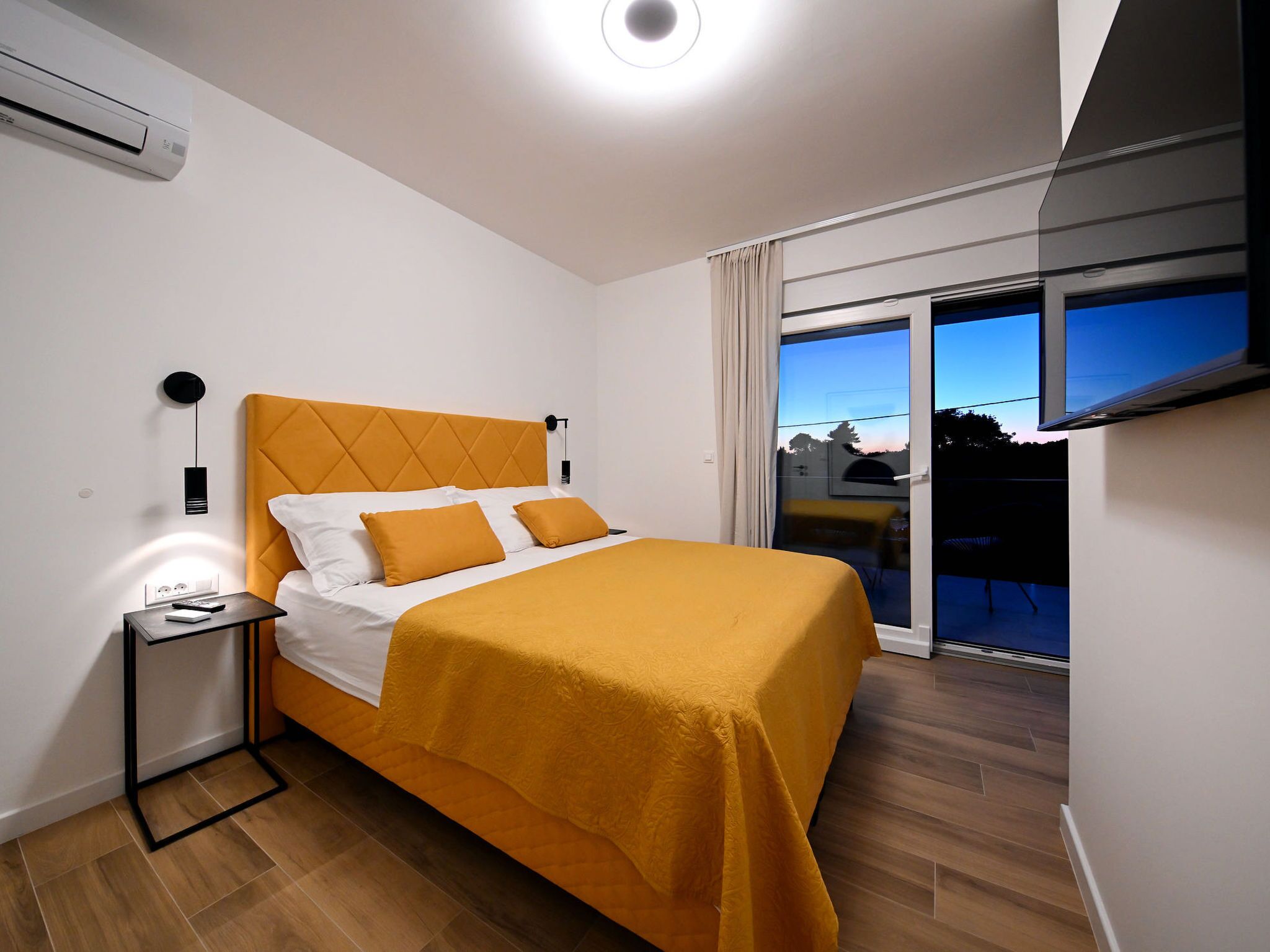 A modern hotel room with a bed, a bedside table, an air conditioning unit, and a large window with a view.