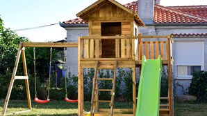 Une maisonnette en bois avec un toboggan et des balançoires, située devant une maison dont le toit est recouvert de tuiles.
