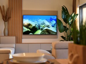 Ein Wohnzimmer mit Fernseher an der Wand, einer Couch, einem Couchtisch und einer Topfpflanze.