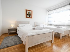 Ein Hotelzimmer mit zwei Betten, einem Nachttisch, einer Lampe, einem gerahmten Bild an der Wand und einem Fenster mit durchsichtigen Vorhängen.