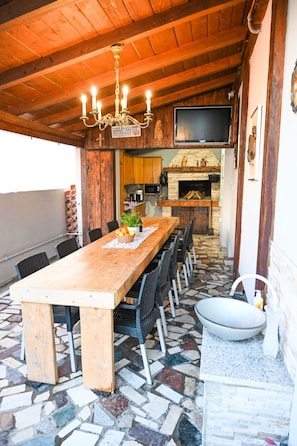 Ein Außenbereich mit Essplatz, einem langen Holztisch, mehreren Stühlen, einem steinbelagten Boden, einem Steinsink und einem Kamin im Hintergrund.
