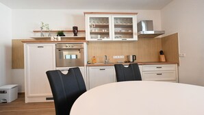 Ett kompakt kök med kylskåp, ugn och skåp, ett matbord med stolar och en liten papperskorg.
