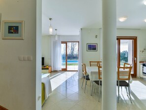 A spacious living area with a dining table, chairs, a TV, and a view of a swimming pool through a glass door. - Vacation home  by Interhome (Privlaka)