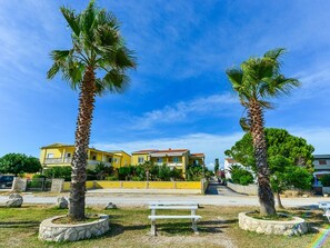 A sunny outdoor area with palm trees, a bench, and a yellow building with a red roof. - Holiday Apartment Inga Blue with exit to the beach by Interhome (Vir)