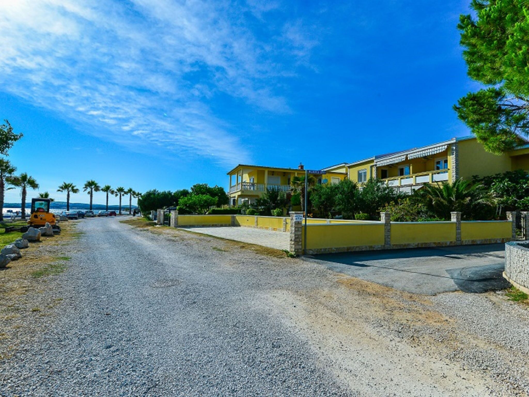 A coastal road leading to a yellow building with a balcony, surrounded by palm trees and a clear sky.