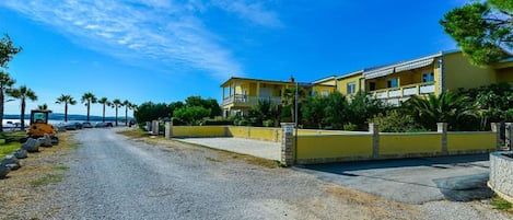 A coastal road leading to a yellow building with a balcony, surrounded by palm trees and a clear sky.