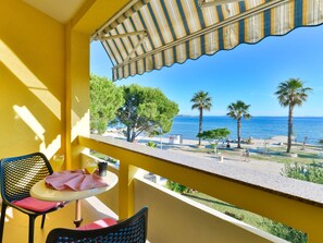 A balcony with yellow walls, a table with a chair, and a view of the sea with palm trees. - Apartment  by Interhome (Vir)