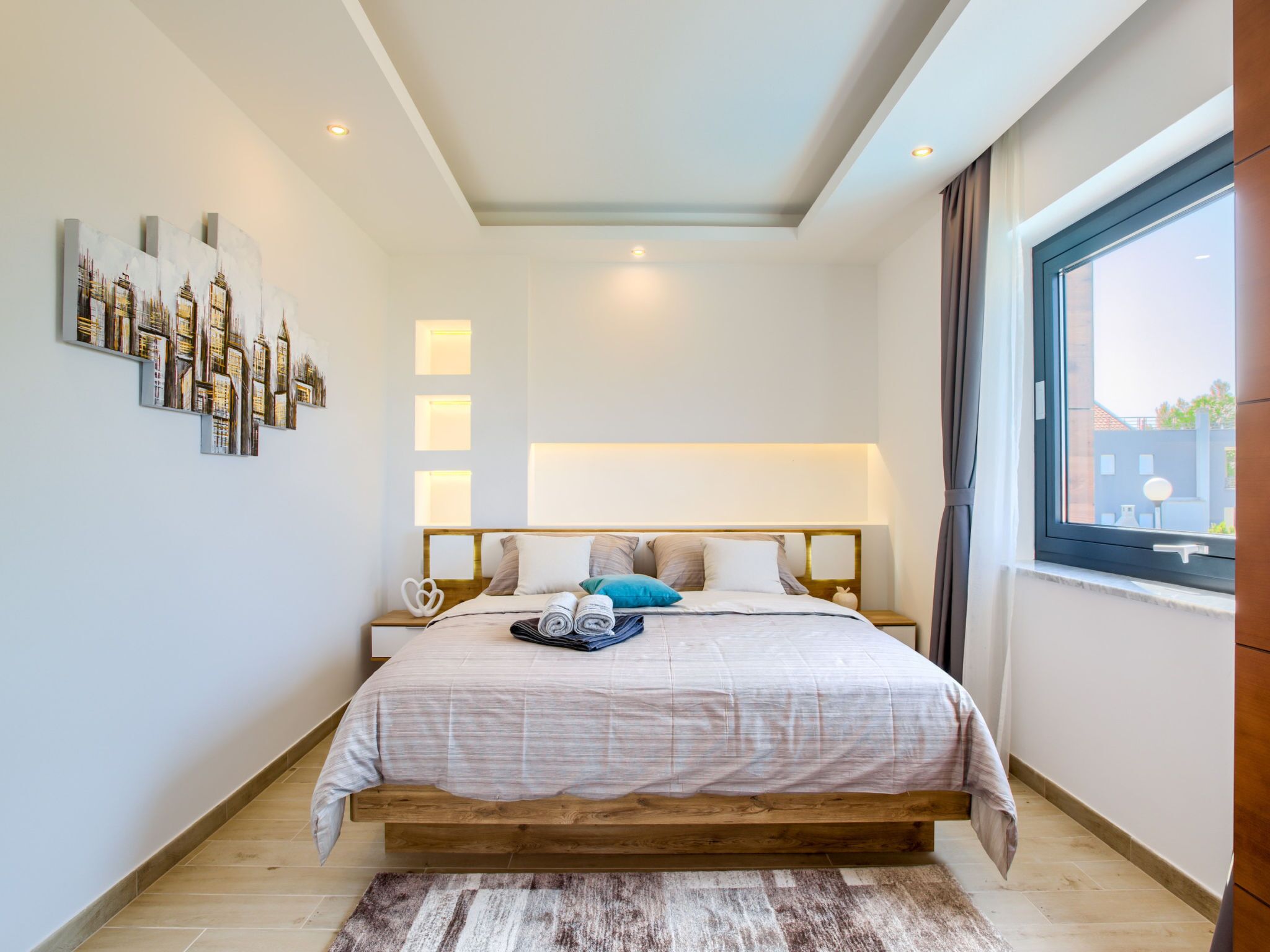 A modern bedroom with a wooden bed, a cityscape painting, recessed ceiling lights, and a large window with curtains.