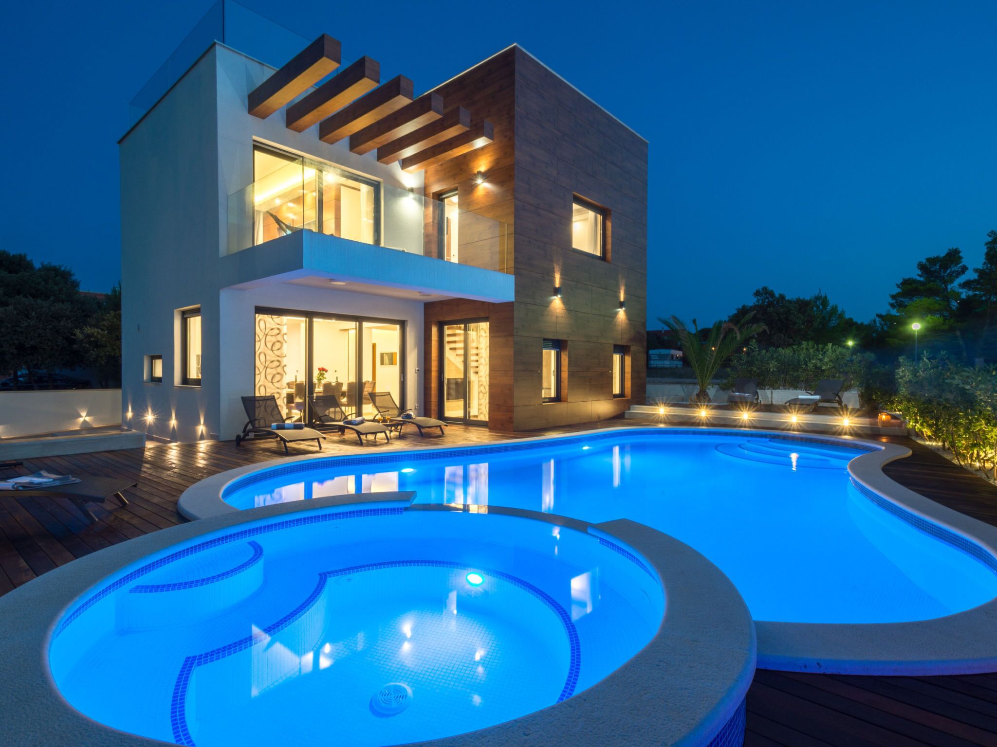 A modern two-story house with a swimming pool, outdoor seating, and a well-lit patio area.