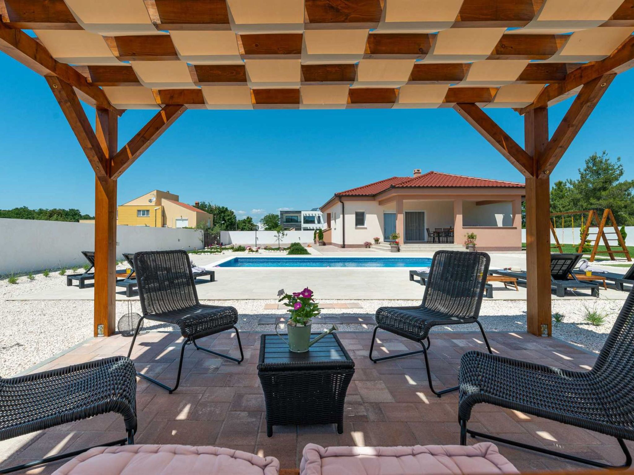 A covered outdoor seating area with wicker chairs and a table, overlooking a swimming pool and a building.