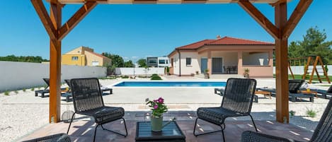 A covered outdoor seating area with wicker chairs and a table, overlooking a swimming pool and a building.