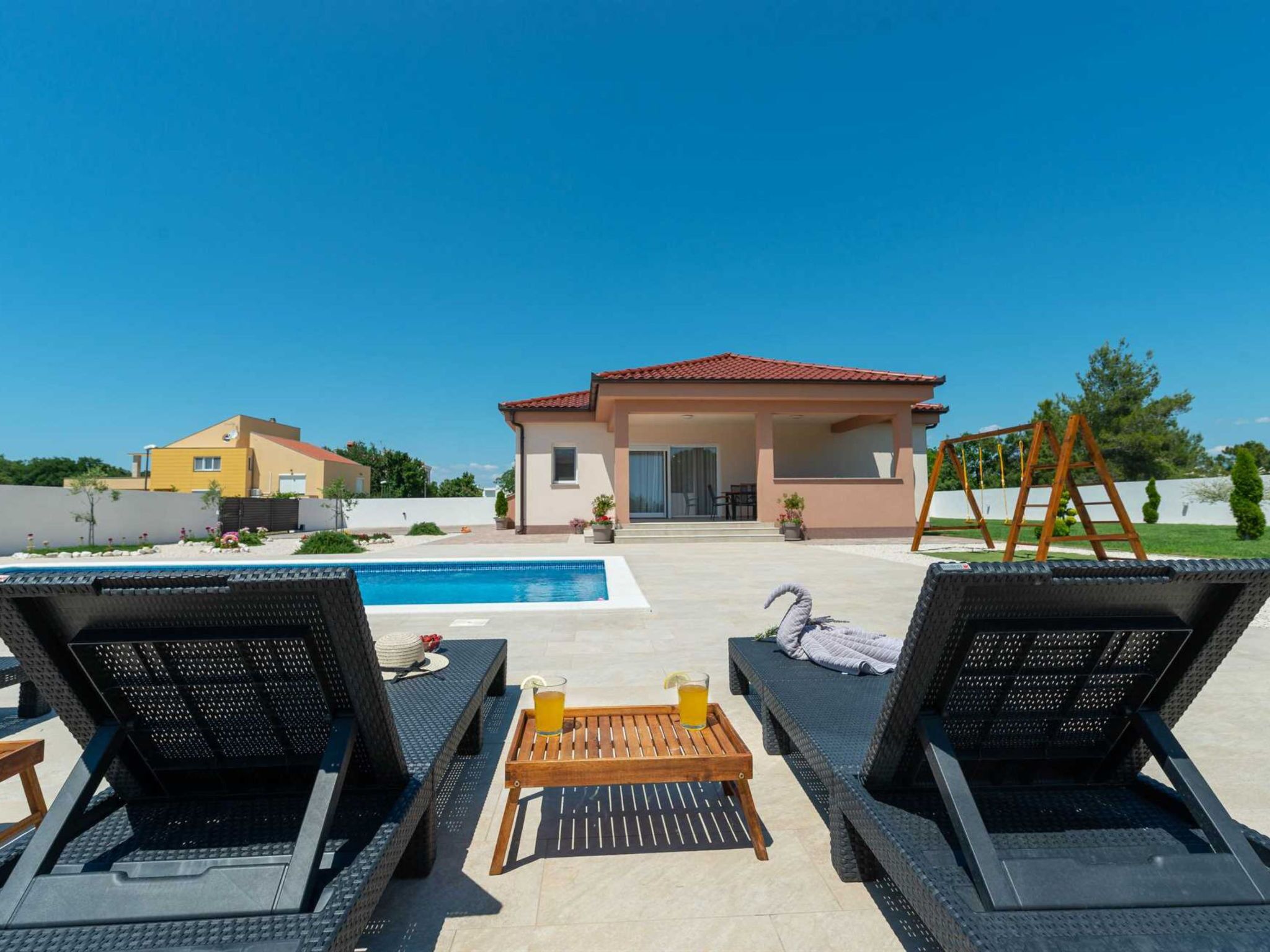 A poolside area with lounge chairs, a wooden table with drinks, and a view of a building with a red roof.