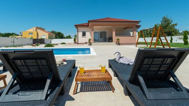 A poolside area with lounge chairs, a wooden table with drinks, and a view of a building with a red roof. - Vacation home  by Interhome (Nin)