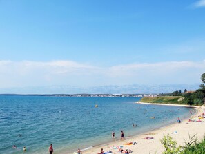 Ein Strand mit badenden und sonnenbadenden Menschen, ein klarer Himmel und Berge im Hintergrund.