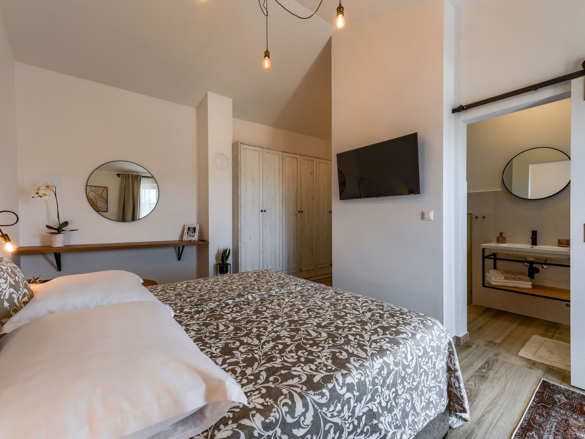 Een slaapkamer met een bed, nachtkastje met lamp, spiegel en een aan de muur bevestigde televisie.