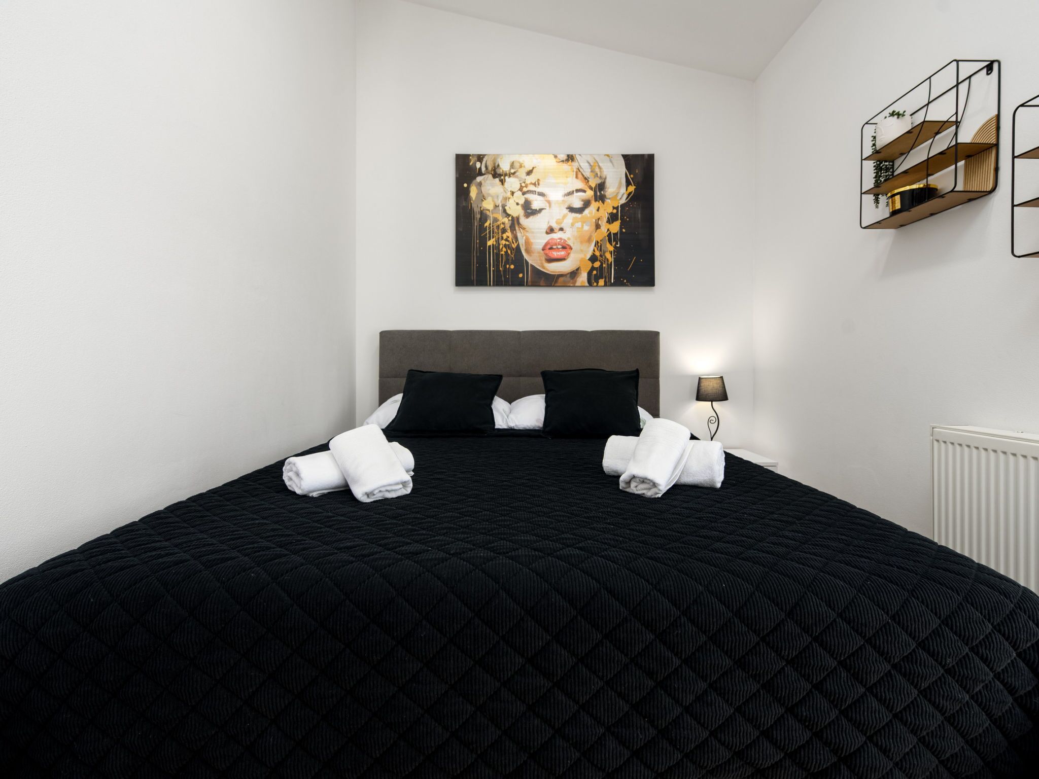Un lit bien fait, avec une literie noire et des serviettes blanches, une tête de lit grise, un tableau au-dessus du lit et une petite étagère avec quelques objets accrochés au mur.