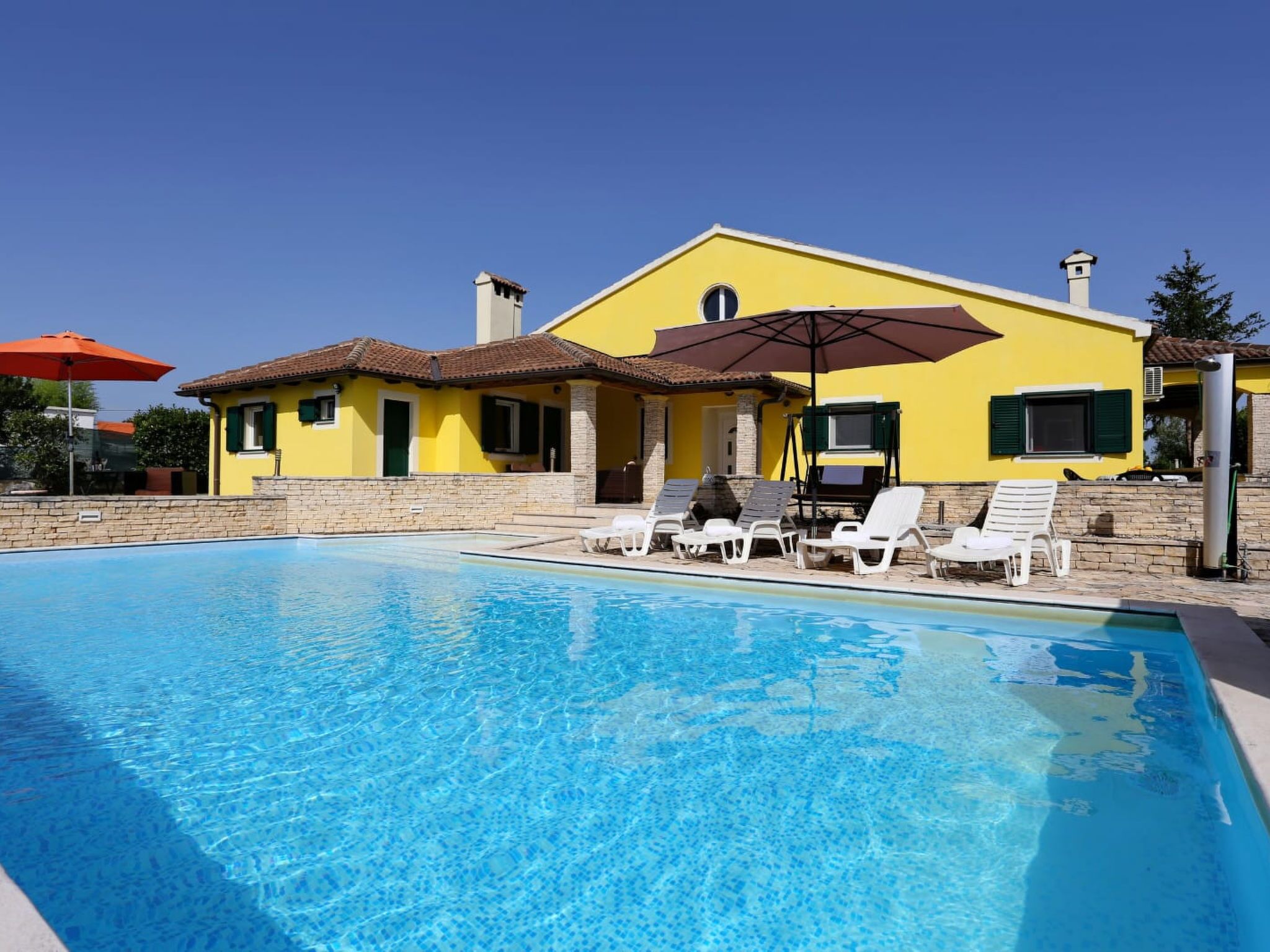 Une piscine avec des chaises longues et des parasols, un bâtiment jaune avec un toit en tuiles et un ciel bleu clair.